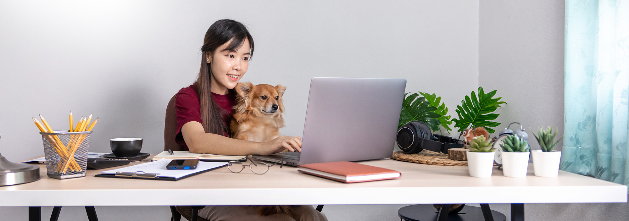 Woman sitting at a desk with a laptop, holding a small dog, in a home office setting.